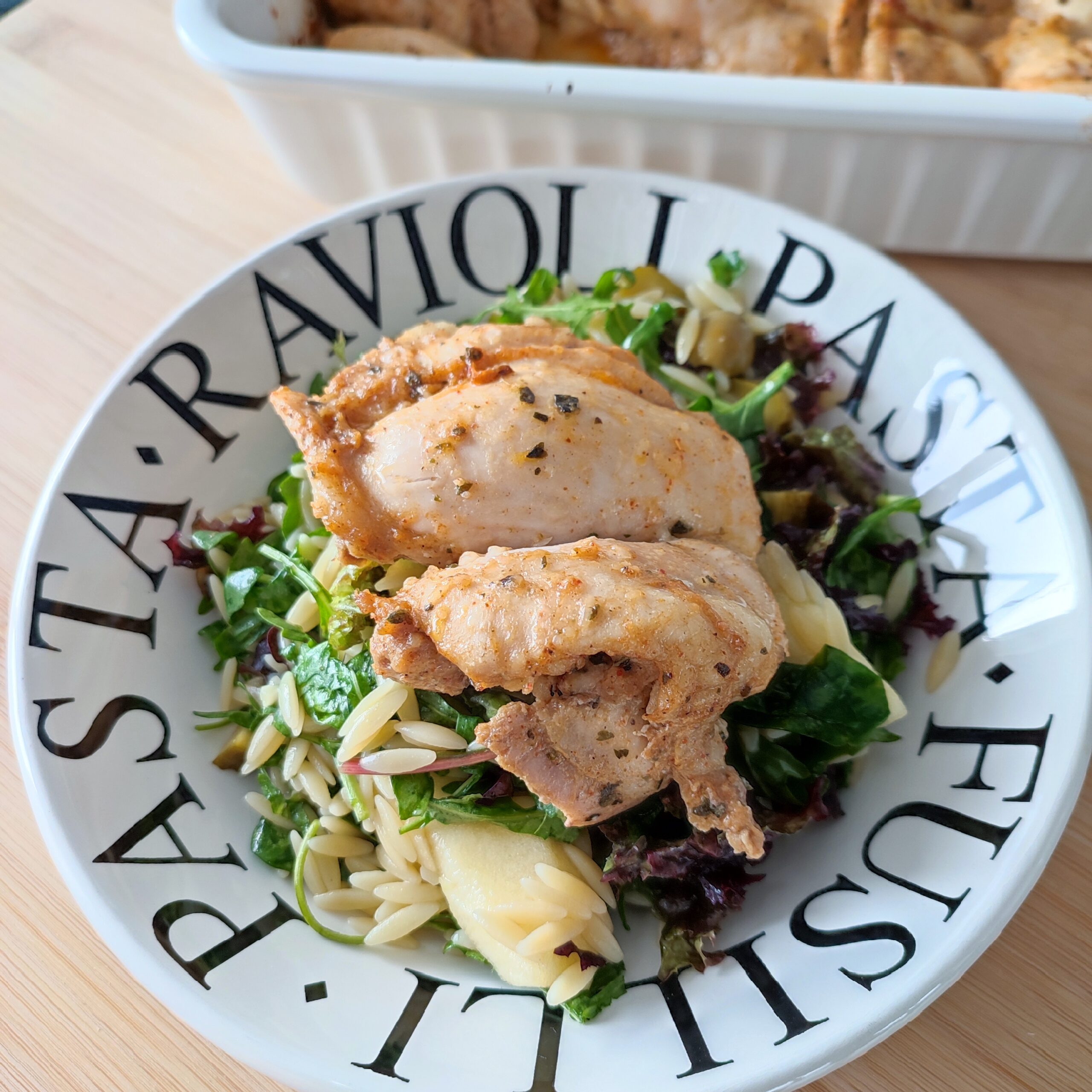 Baked Parmesan Chicken with the eveyrthing salad (orzo added) in a pasta serving plate on a working surface and the casserole dish with the remaining baked parmesan chicken in the background