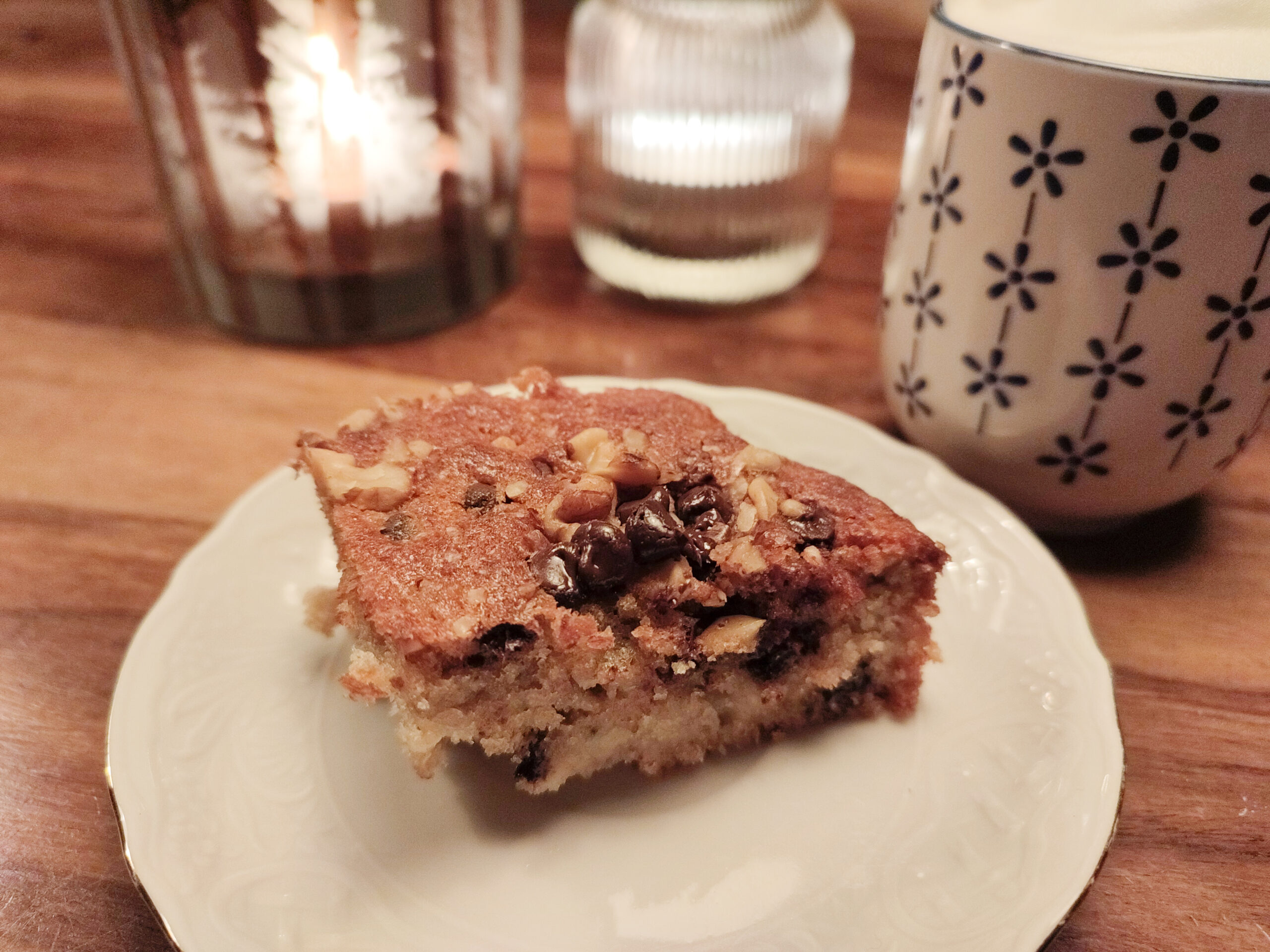 Slice of banana walnut chocolate cake on a white plate with latte in a blue cup and candles in fancy glassware