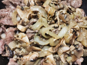 Adding sauteed mushrooms, garlic and onion to beef in a pan