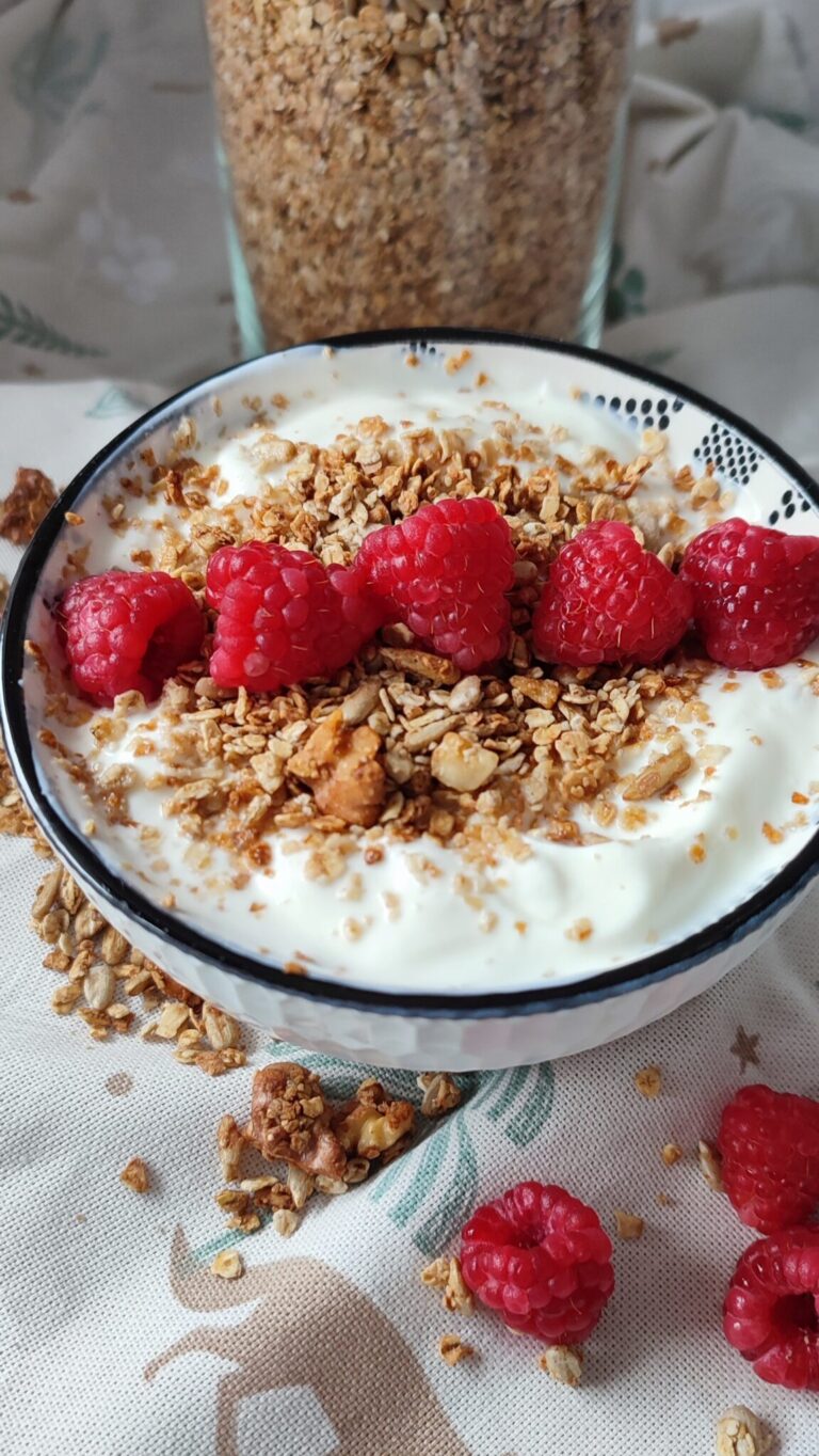 Yoghurt in a small bowl topped with granola and fresh raspberries with the stored granola in container in the background