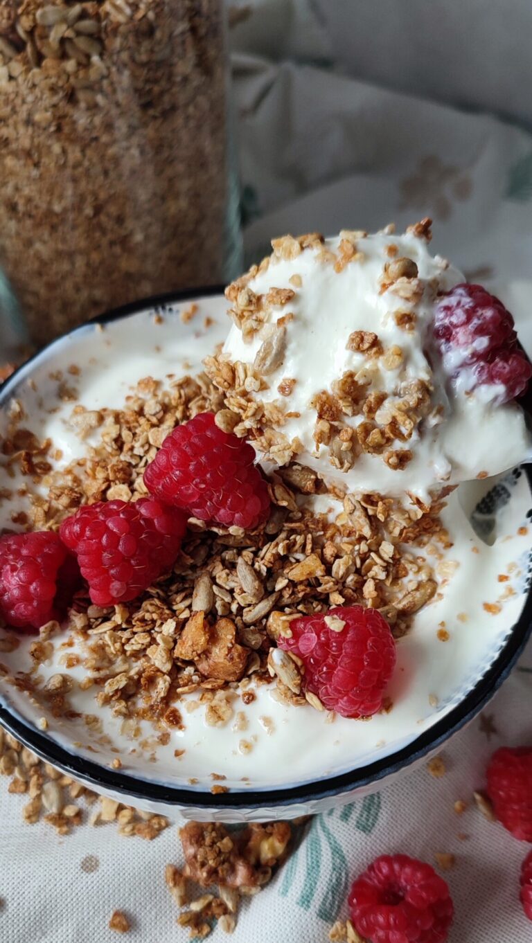 Yoghurt in a small bowl topped with granola and fresh raspberries with the stored granola in container in the background