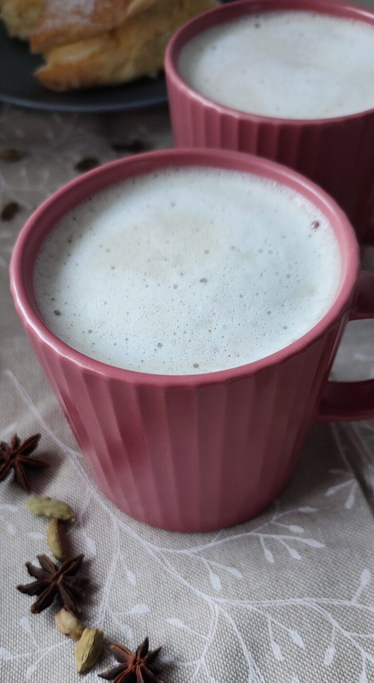 Masala Chai Latte accompanied by another cup of masala chai latte, trail of spices and cream buns in the background