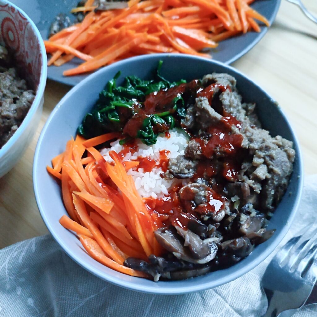 Bulgogi Bibimbap in a bowl with ingredients in the background and cutlery beside the bowl on a patterned fabric