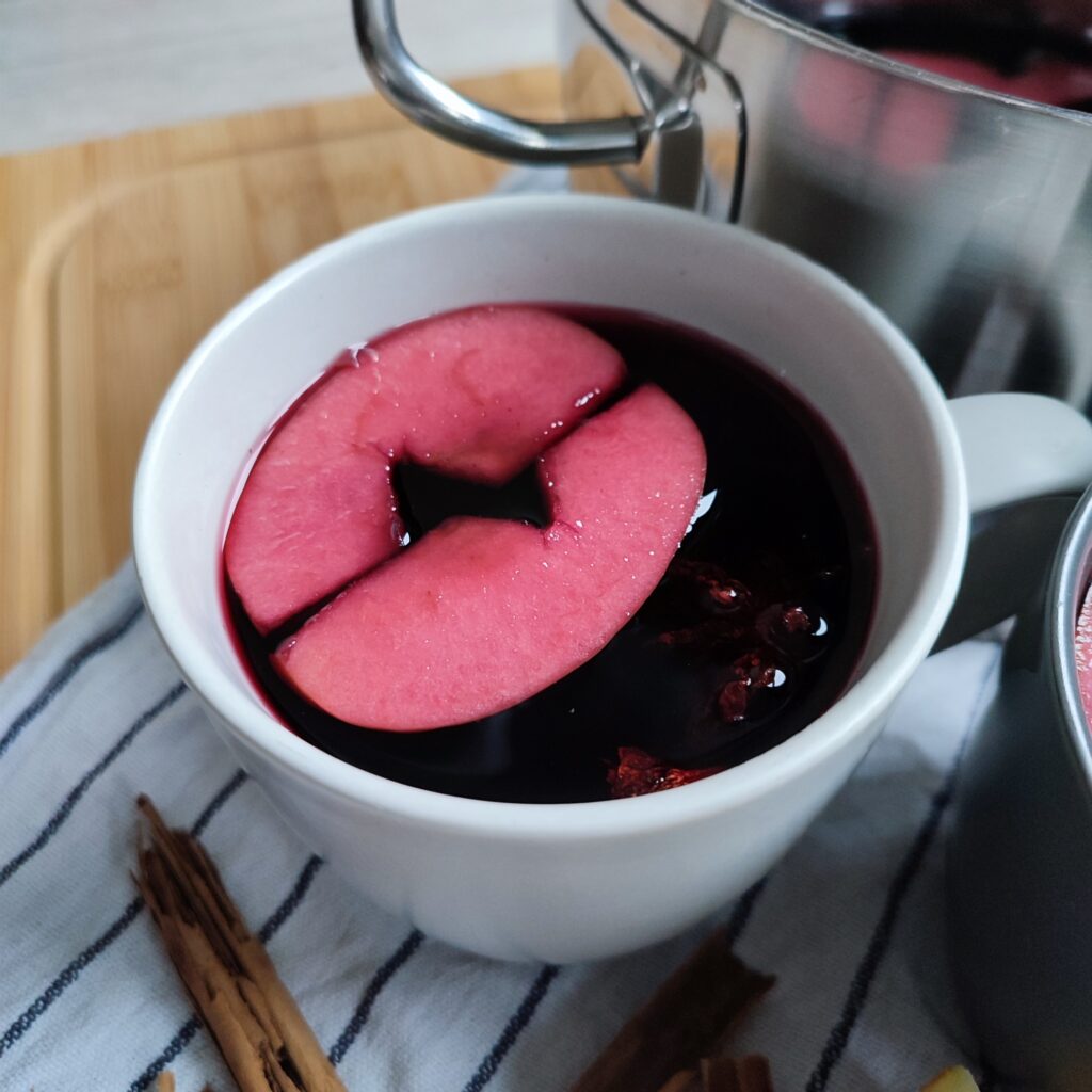 A full cup of Glühwein, with apple pieces floating in it and a pot of Glühwein can be seen in the background. Cinnamon sticks are scattered around on a striped cloth atop a working surface, serving as decoration.