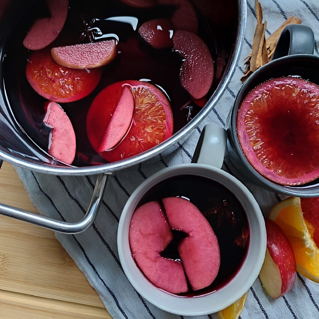 A pot filled with Glühwein sits beside two full cups of Glühwein, each with fruit pieces floating in them. Cinnamon sticks, orange slices, and apple slices are scattered around on a striped cloth atop a working surface, serving as decoration.