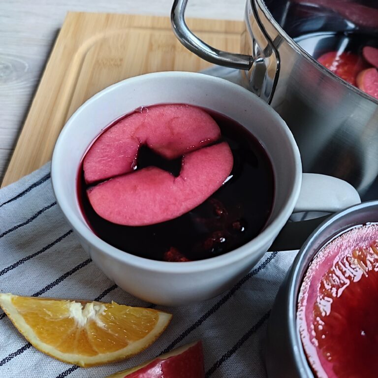 A full cup of Glühwein, with apple pieces floating in it and a pot of Glühwein can be seen in the background. Orange slices, and apple slices are scattered around on a striped cloth atop a working surface, serving as decoration.