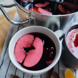 Two full cup of Glühwein, with fruit pieces floating in it and a pot of Glühwein can be seen in the background.