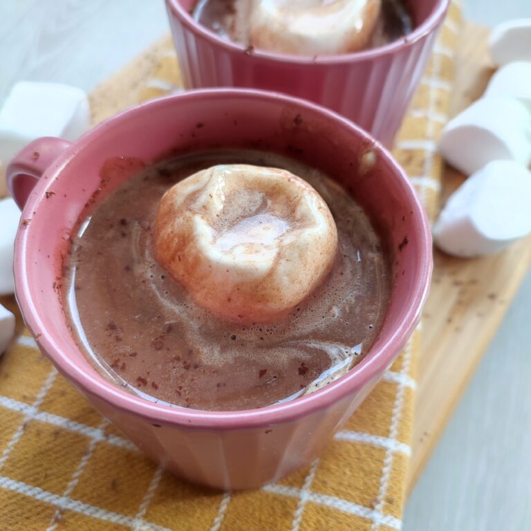 Creamy Hot Chocolate with marshmallow and chocolate shavings in a pink cup placed on a patterned yellow cloth on a working surface decorated with some marshmallows on the side