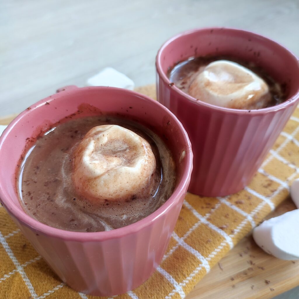 Creamy Hot Chocolate b - roll 1 : Hot Chocolate with marshmallow and chocolate shavings in two pink cups placed on a patterned yellow cloth on a working surface decorated with some marshmallows on the side