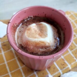 Hot Chocolate with a marshmallow in it on a yellow patterned cloth with a few marshmallows as decoration