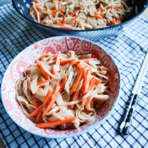 A side angle of quick stir fry yakisoba in a wok and a portion of stir Fry Yakisoba in a decorative bowl on a checkered cloth with a pair of chopsticks on the side