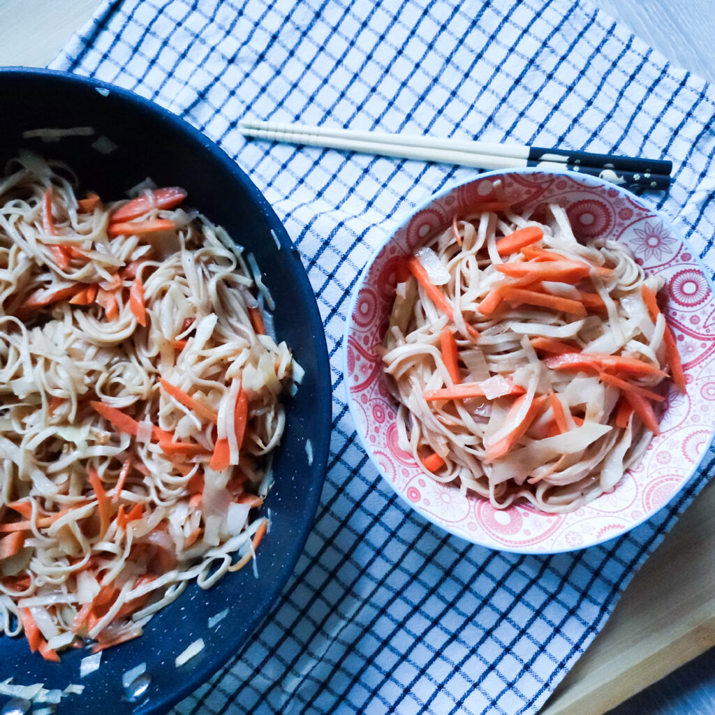 An upper angle of quick stir fry yakisoba in a wok and a portion of stir Fry Yakisoba in a decorative bowl on a checkered cloth with a pair of chopsticks on the side