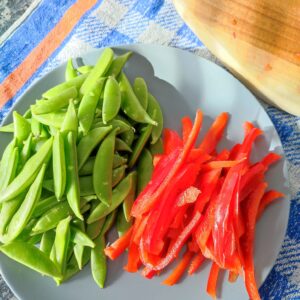 Sugar snap peas and thinly sliced bell pepper on a blue plate with a chopping board beside it