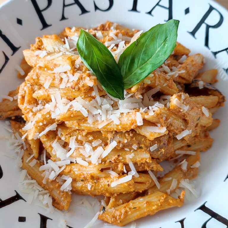 Pasta Pesto Rosso with Shiitake Mushrooms garnished with shredded parmigiano reggiano and basil leaves on a white plate with black lettering