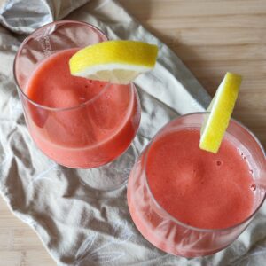 Pink Sgroppino in wine glasses garnished with quartered lemon wedges on the side on patterned taupe coloured fabric on a working surface