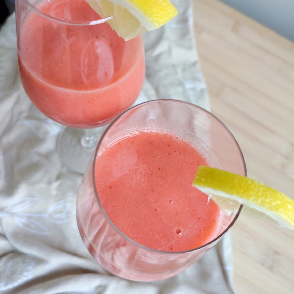 B roll 2: Pink Sgropinno in two wine glasses with a wedge of quartered lemon on the rims standing on taupe coloured fabric on a working surface top shot