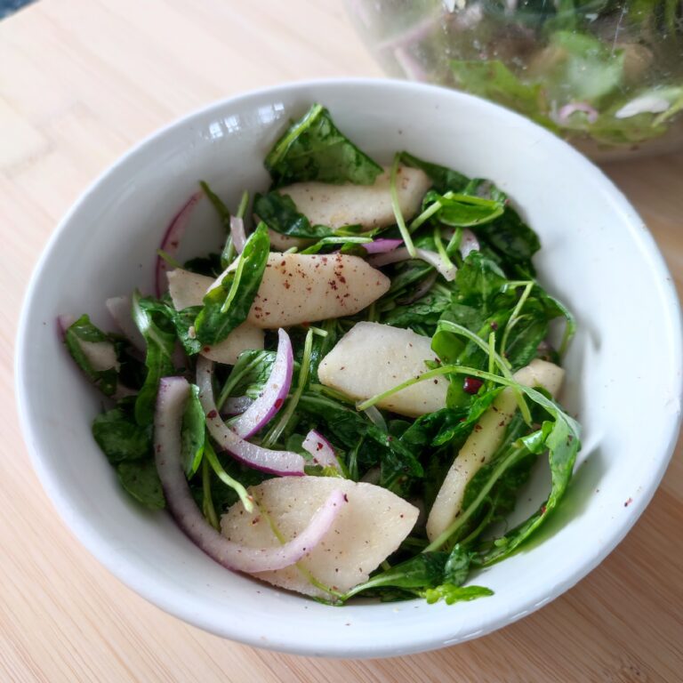 Apple Arugula Sumac Salad in a white salad bowl on a working surface with a large clear bowl in the background with more salad in