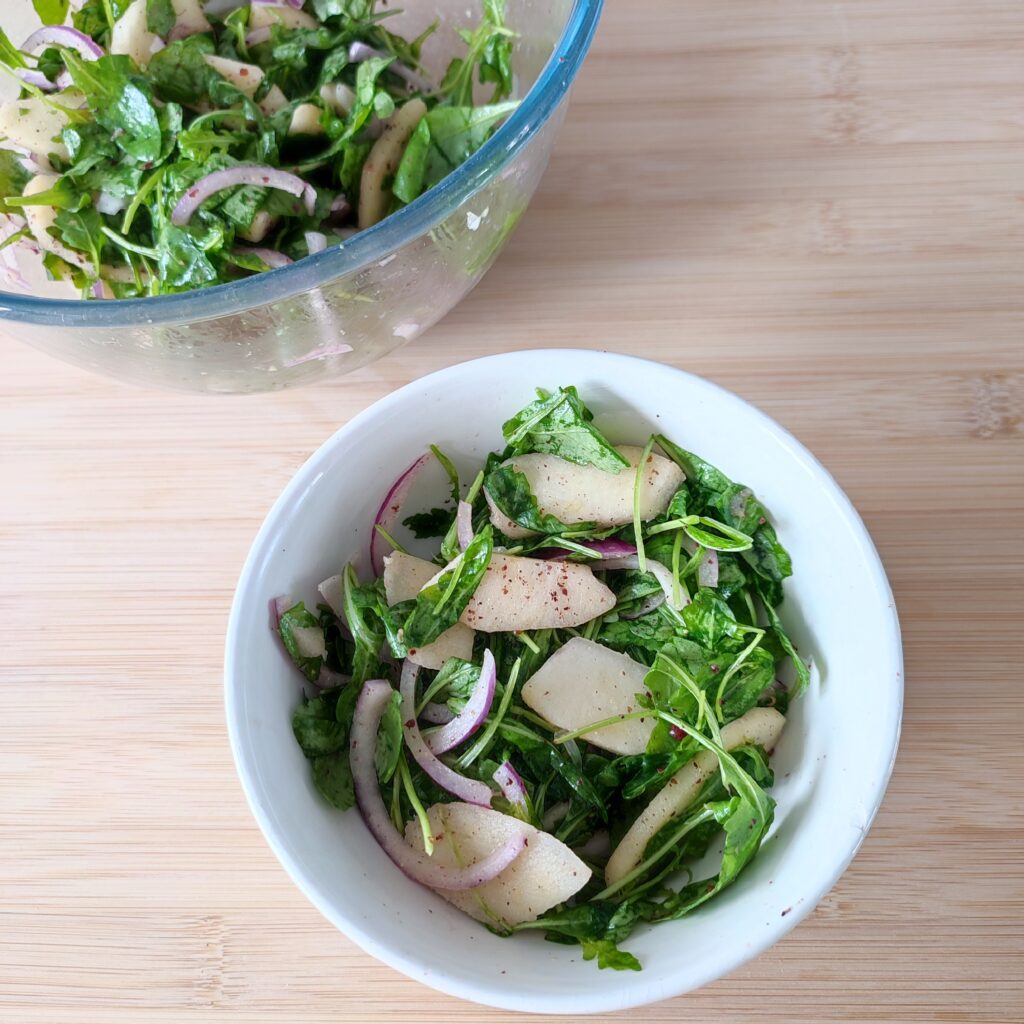 B roll 1: Apple Arugula Sumac Salad in a white salad bowl on the wroking surface with a large clear bowl with remaining salad beside it