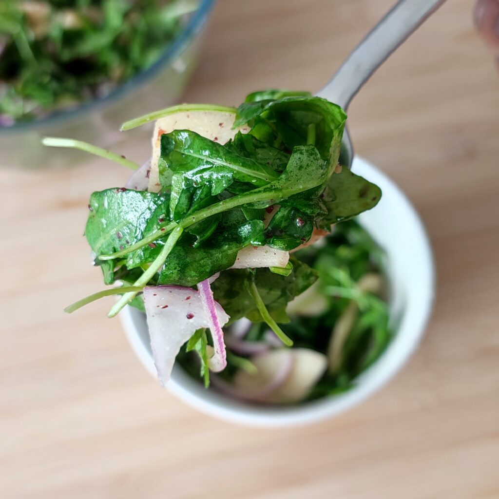 B roll 2: A forkful of Apple Arugula Sumac Salad on a fork with a serving of salad in a white salad bowl and a large clear bowl with the remaining salad in the background on a working surface