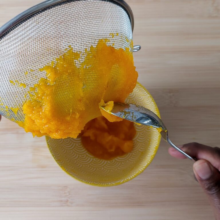 Scraping the pureed pumpkin from the strainer' s bottom using a spoon with the small yellow patterned bowl with some of the pumpkin puree in the background