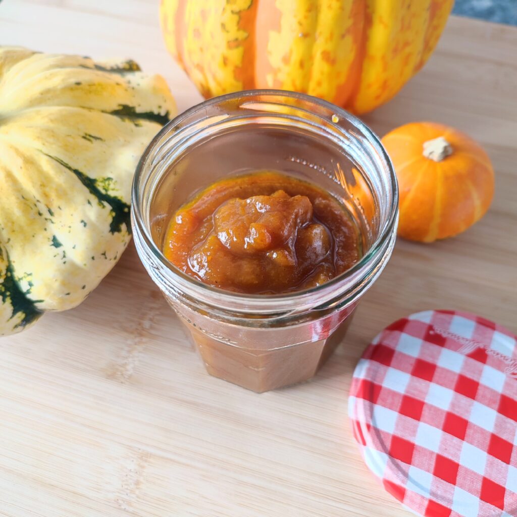 B roll 1: Pumpkin Spice Syrup in a small confiture jar with its red and white checkered patterned lid resting on the side flat horizontally and three decorative pumpkins in the background