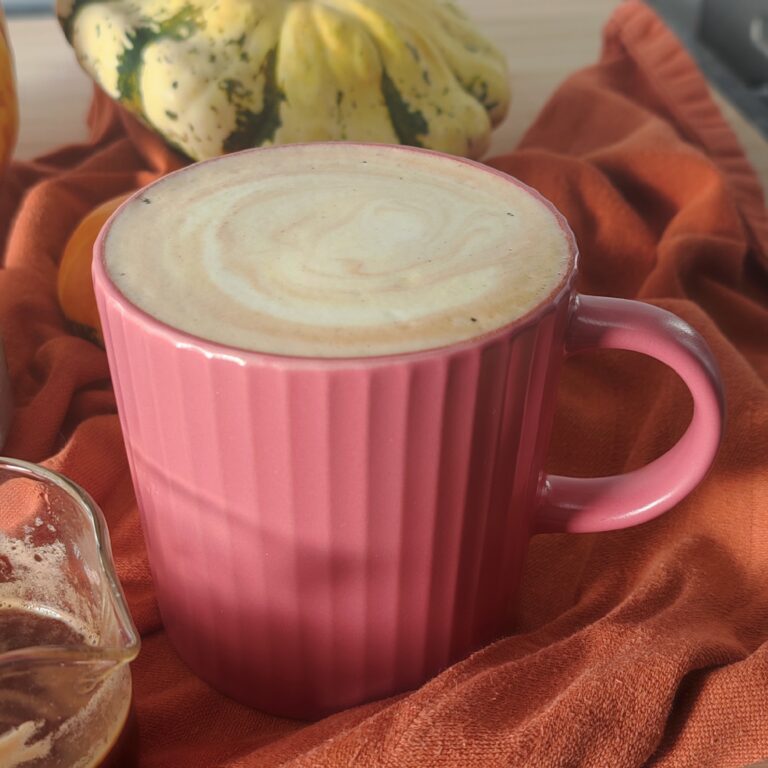 Dirty Pumpkin Spice London Fog Latte accompanied by a shot of espresso in a shot glass with a stainless steel mug and pumpkins on burnt orange fabric