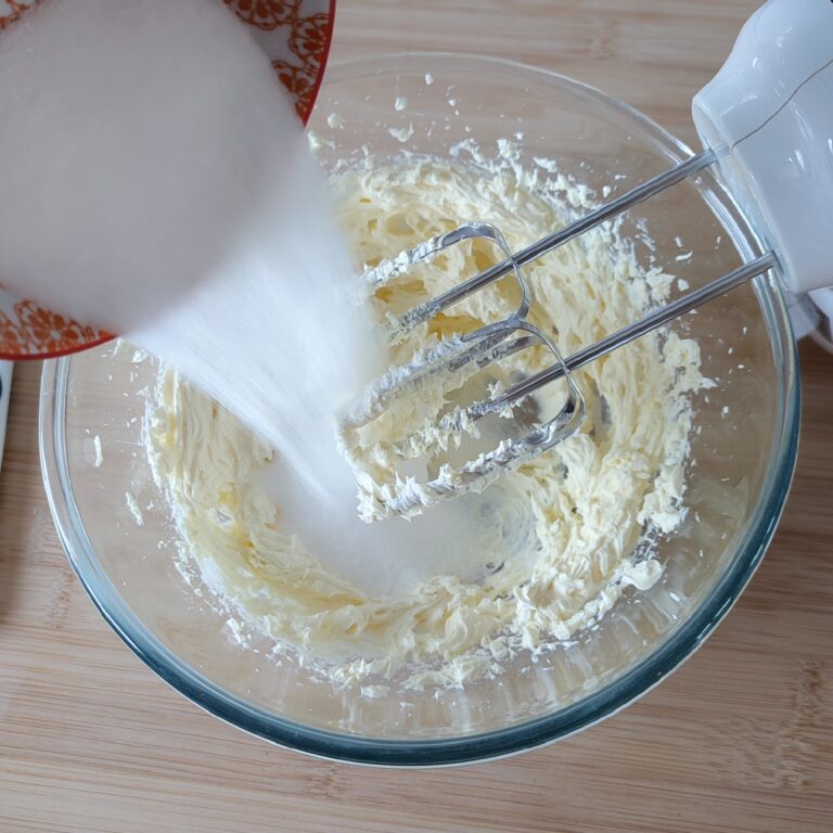 Adding sugar to beaten cream cheese in a large clear bowl