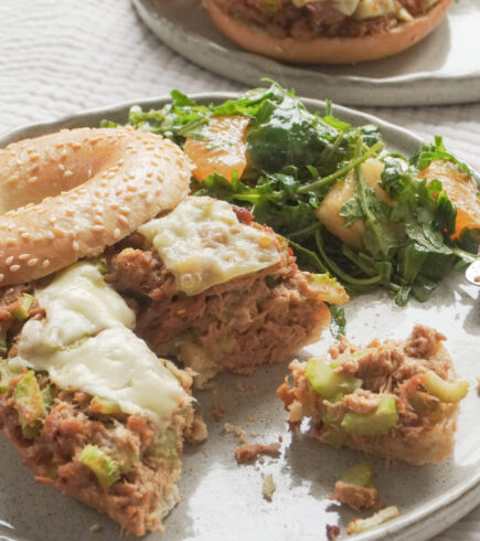 Half eaten Airfryer Tuna Bagel Melt with a side of wild peach and mandarin arugula salad on modern ceramic plate and another airfryer tuna bagel melt in the background on textured beige table cloth