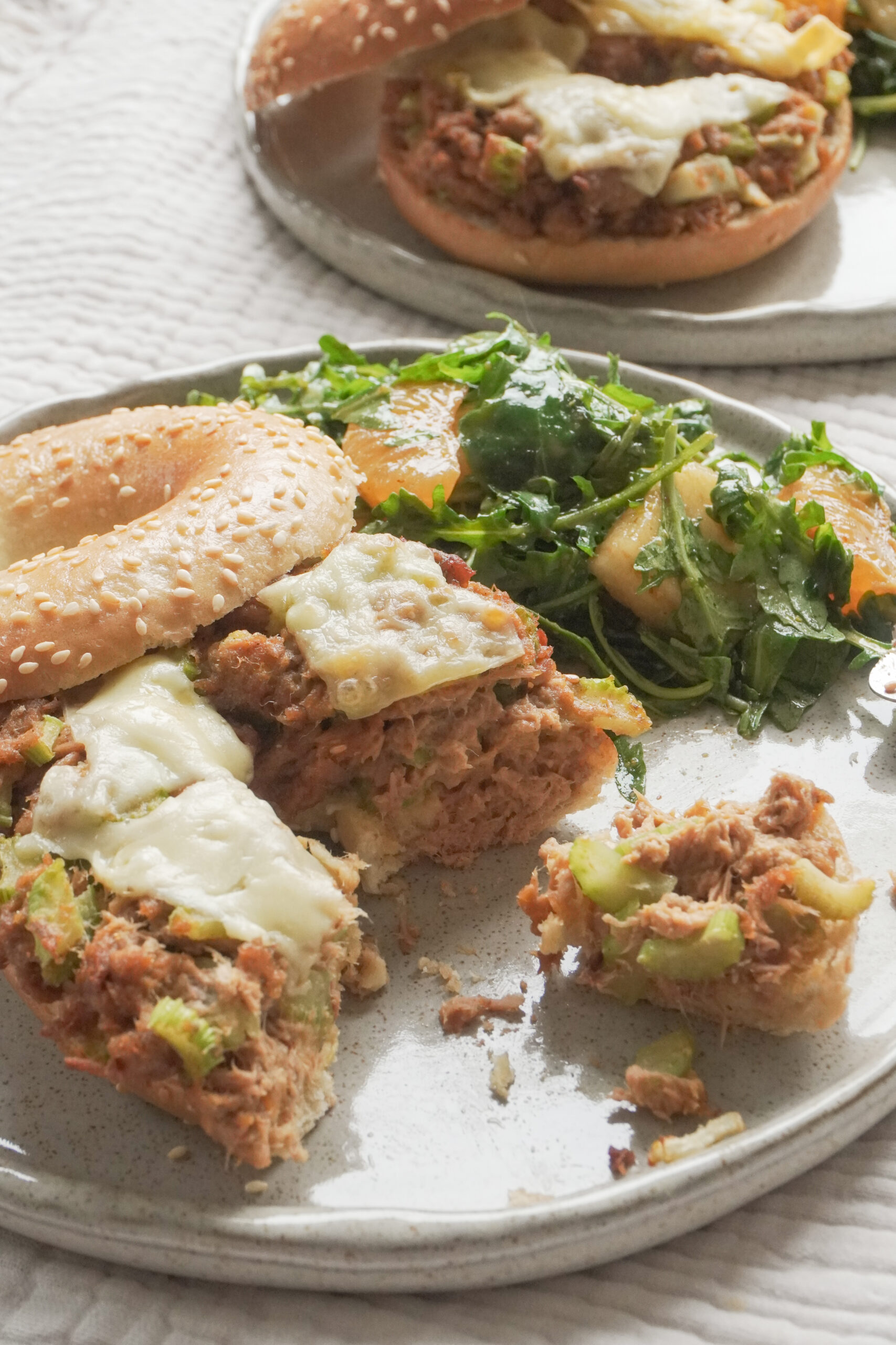 Half eaten Airfryer Tuna Bagel Melt with a side of wild peach and mandarin arugula salad on modern ceramic plate and another airfryer tuna bagel melt in the background on textured beige table cloth