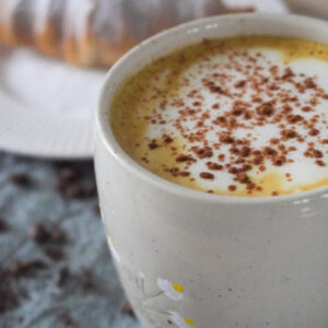 Spiced Pear & Turmeric Maple Latte with Soy Milk in a beige mug with a croissant with powdered sugar dusted on it on a white plate in the background and both are on a green cloth with spices, coffee beans, matcha powder and tea leaves scattered