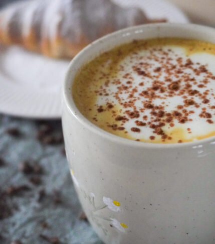 Spiced Pear & Turmeric Maple Latte with Soy Milk in a beige mug with a croissant with powdered sugar dusted on it on a white plate in the background and both are on a green cloth with spices, coffee beans, matcha powder and tea leaves scattered