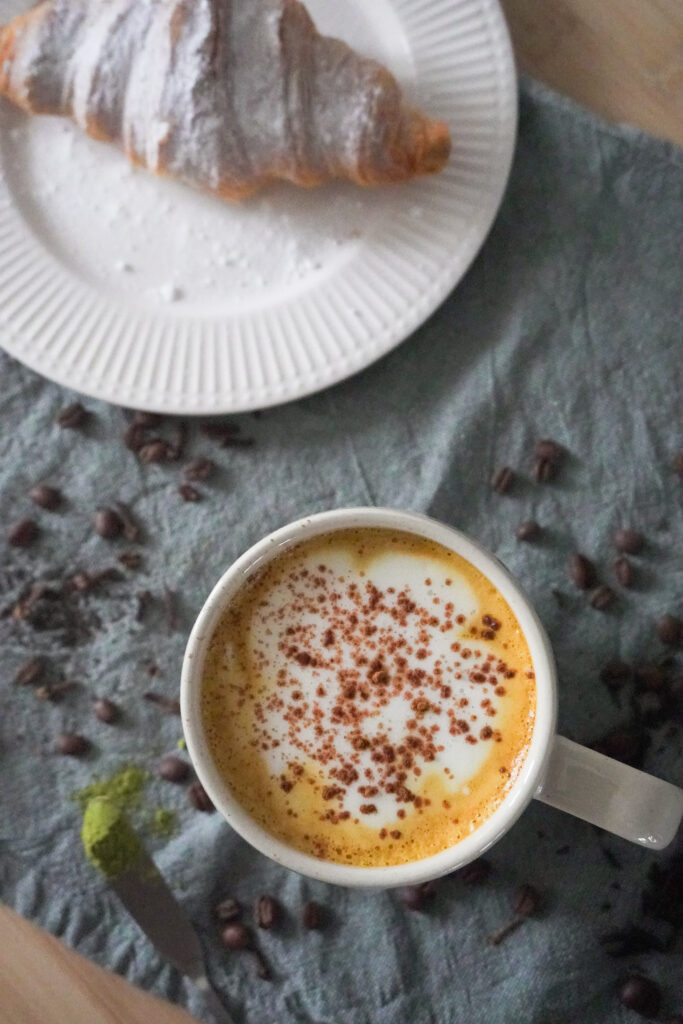 Spiced Pear & Turmeric Maple Latte with Soy Milk in a beige mug on the far right with a croissant with powdered sugar dusted on it on a white plate opposite on the far left and both are on green cloth with spices, coffee beans, matcha powder and tea leaves scattered