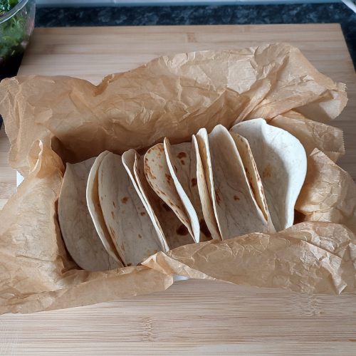 Prepped soft tortillas aligned next to each other in a off white casserole dish that is lined with crinkled up parchment paper on a working surface