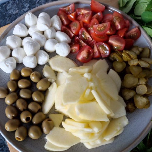 Thin apple slices, quartered cherry tomatoes, sliced gherkins, green olives and mini mozzarella balls on a large blue plate